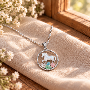 A silver-toned necklace with a circular pendant featuring a white horse and small pastel gemstones, displayed on beige fabric near a wooden window with small white flowers.