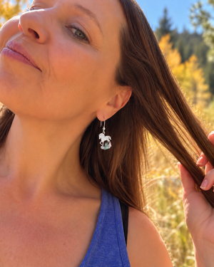 A close-up lifestyle shot of a woman outdoors wearing a silver dangle earring shaped like a white and black dog.