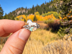 A hand holding a silver ring featuring a white horse silhouette and small pastel gemstones, with a blurred outdoor mountain landscape in the background.