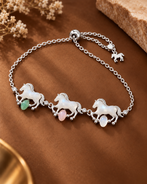 A silver-tone chain bracelet featuring three running horse charms with white mother-of-pearl details and small dangling green and pink stones, displayed on a brown satin-like surface with dried flowers and a stone prop.