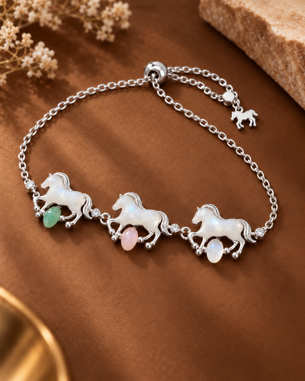 A silver-tone chain bracelet featuring three running horse charms with white mother-of-pearl details and small dangling green and pink stones, displayed on a brown satin-like surface with dried flowers and a stone prop.