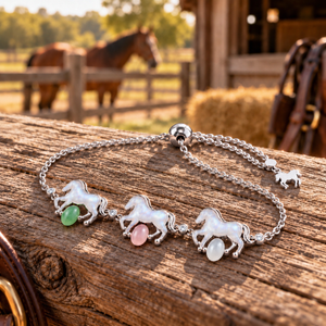 A silver horse charm bracelet with three small horses and green, pink, and white bead accents, displayed on a rustic wooden log with a blurred barn and horse in the background.