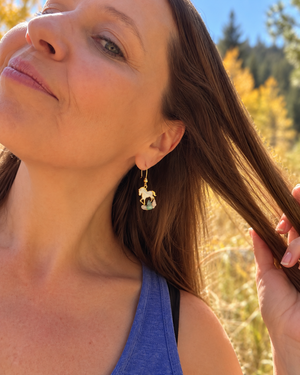 A close-up lifestyle shot of a woman outdoors wearing a gold dangle earring with a small white and green horse charm, set against a blurred background of golden grass and trees.