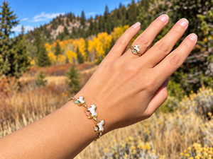 A person's hand wearing a gold bracelet and matching ring, both featuring white dog-shaped charms with colorful gemstone accents, set against a blurred autumn forest background.