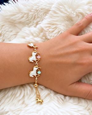 A gold-toned charm bracelet with three white unicorn charms, small pastel pink and mint green beads, and a dangling gold unicorn charm, shown worn on a person's wrist over a white faux fur background.