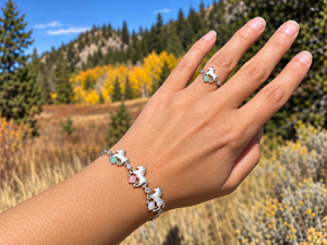 A hand wearing a silver charm bracelet and matching ring with white horse charms and small pastel gemstones, photographed outdoors against a blurred mountain and autumn forest background.