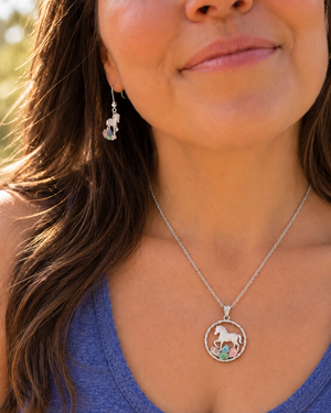 A close-up lifestyle shot of a woman wearing a gold necklace with a circular pendant featuring a white horse and small green and blue accents, along with a matching dangling earring.