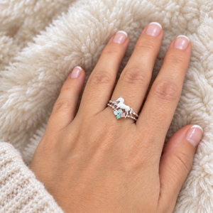 A close-up of a hand wearing a silver ring with a unicorn design and small light blue flower accents, resting on a soft cream-colored faux fur surface.